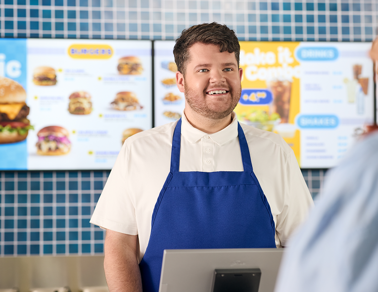 Two people stand at a counter of a quick service restaurant with their backs toward the camera as they look at the vibrant offering on an overhead indoor digital menu board.