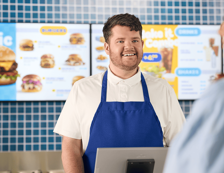 Two people stand at a counter of a quick service restaurant with their backs toward the camera as they look at the vibrant offering on an overhead indoor digital menu board.