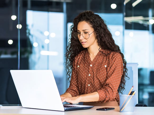 A person with long, dark hair wearing glasses and a rust-colored, polka-dot shirt sits in an office using a laptop computer