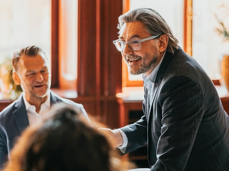 A man leans over a conference table and smiles as he speaks to other gathered there.
