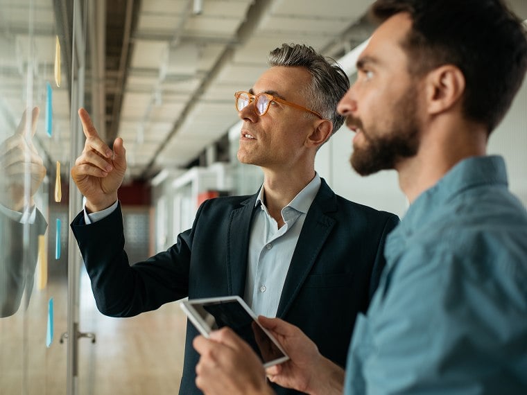 Two men stand in front of a plexiglass board with sticky notes on it as they discuss business.