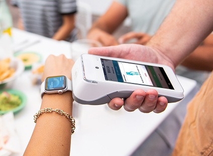 A closeup of a person tapping their smart watch to a mobile payments device to complete a NFC transaction.