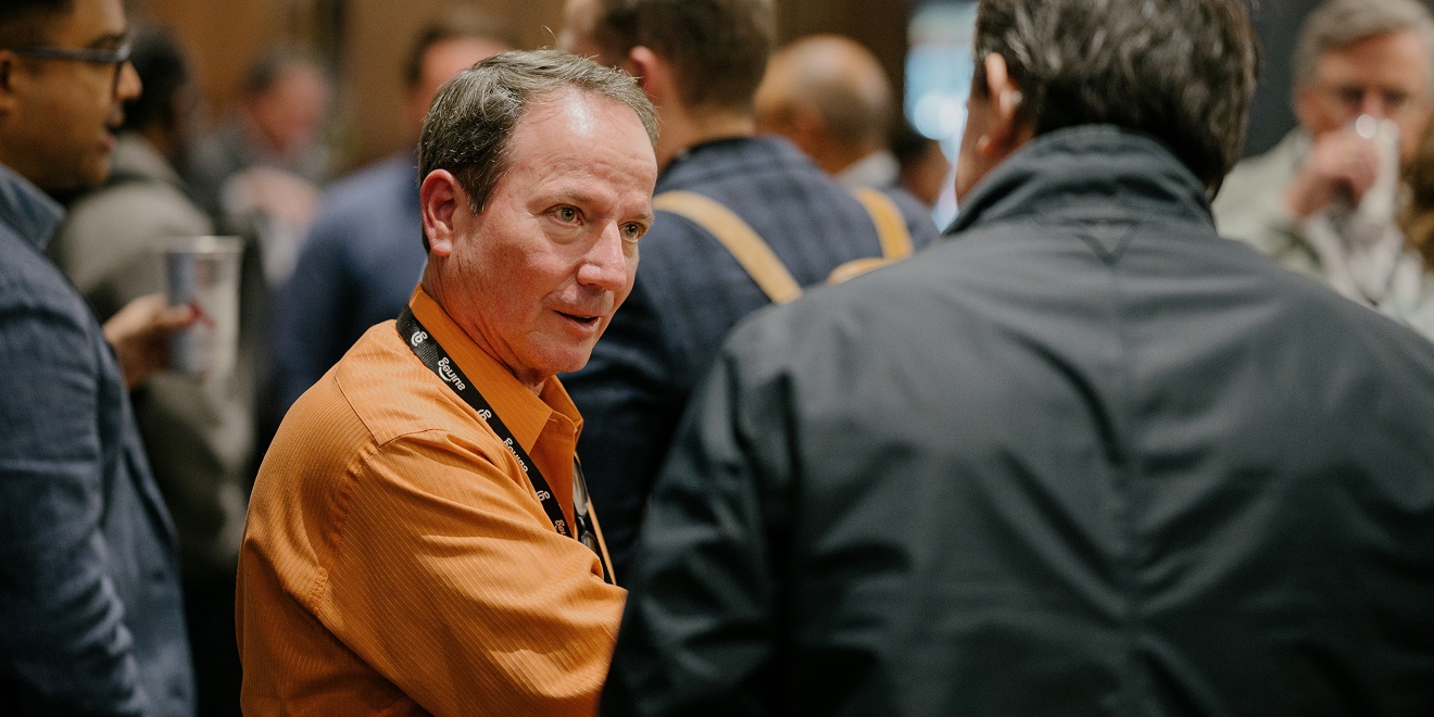 A man wearing a lanyard speaks to another man in a crown at a ISO tradeshow event.