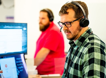 An employee wears an over the ear headset with a mic as he works in front of some computers.