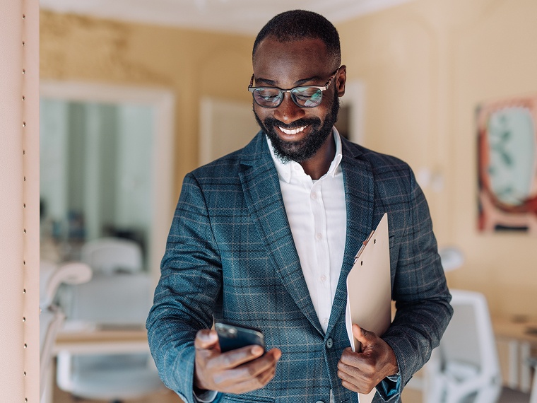 A smiling man in an office setting holds a clipboard under his arm and looks down at the phone in his other hand.