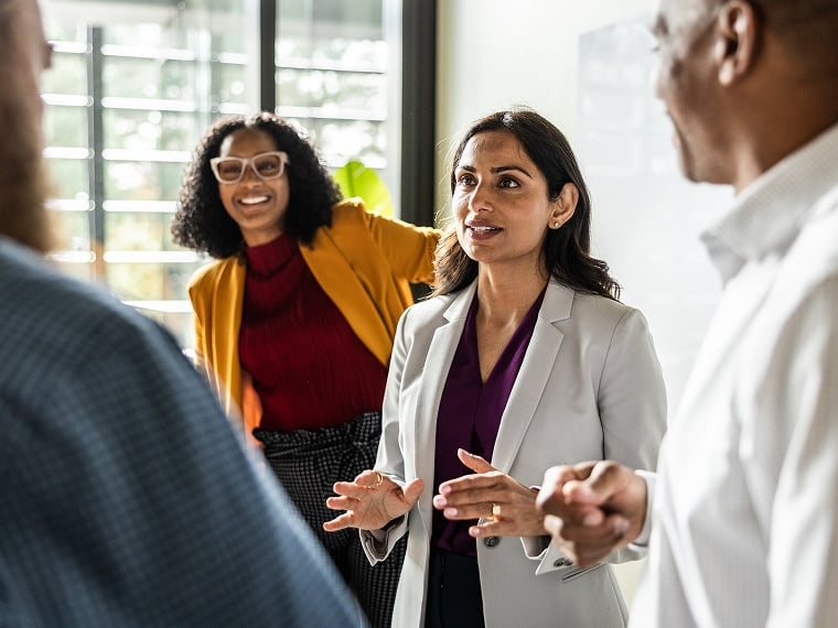 A group of four people stand together in an office building setting while discussing business.