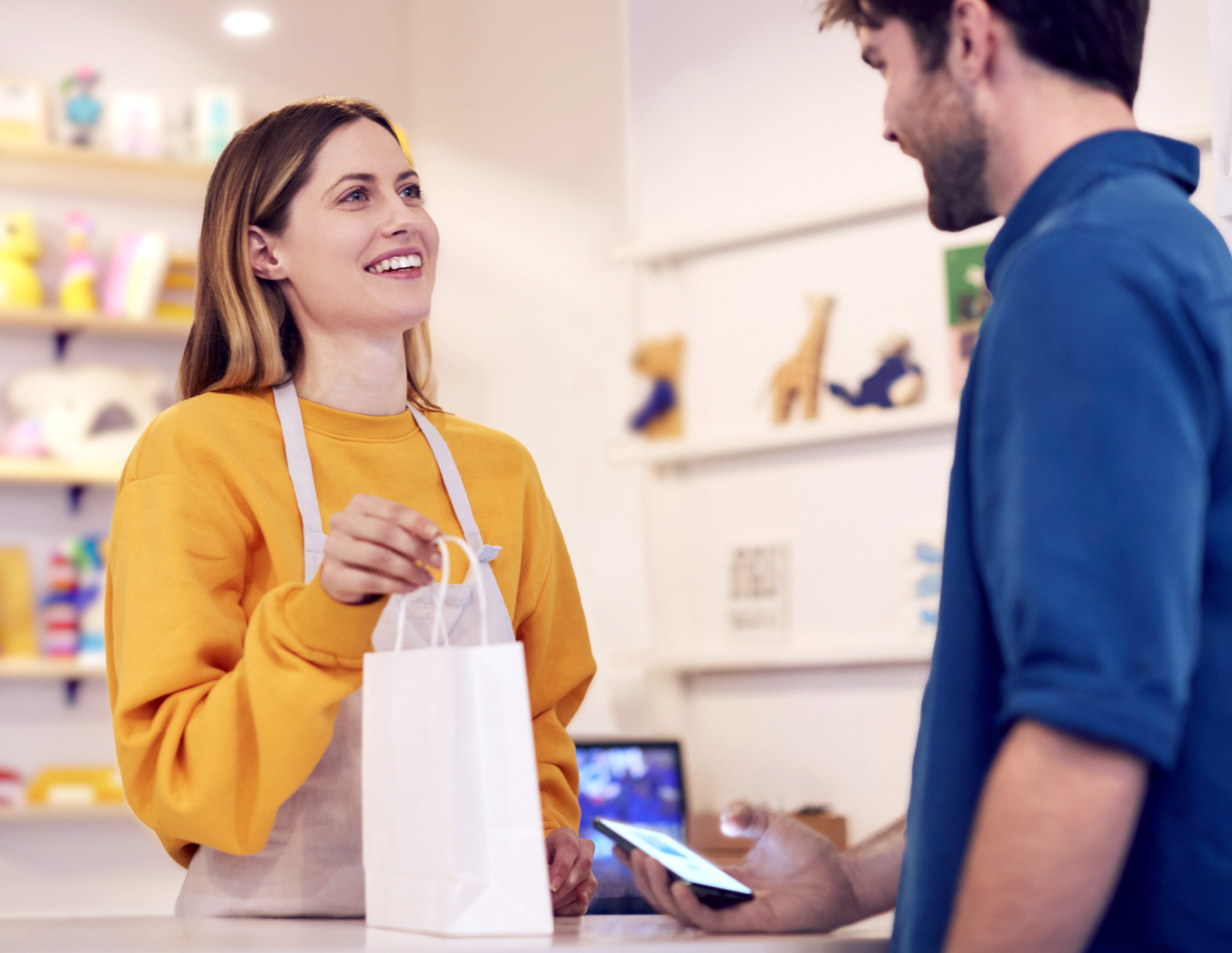 A smiling shopkeeper hands a bag to a customer who’s using a phone to pay for the purchase