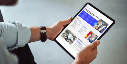 Hands holding a tablet showing a report spread with blue headings, body text, and multiple photos.