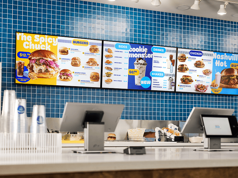 Large digital menu boards sit above the food prep station of a restaurant.