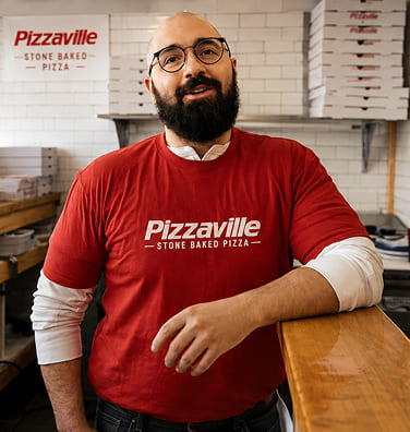 Man in a red Pizzaville shirt leaning on a counter in front of pizza boxes.
