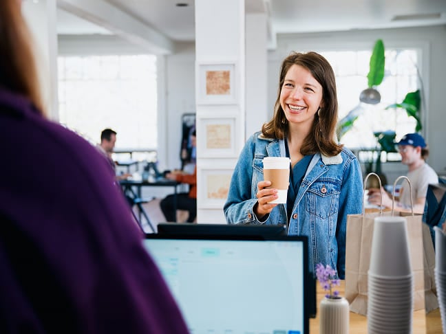 Smiling woman holding a coffee cup at a cafe checkout counter.