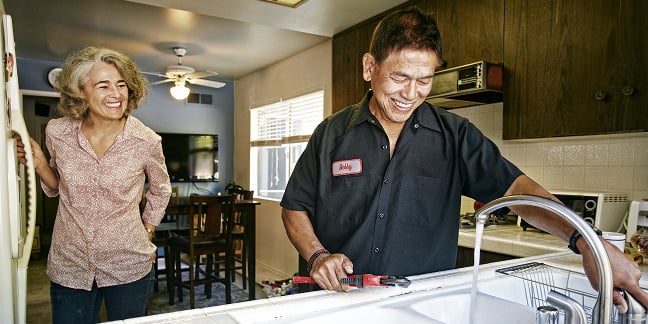 Un homme à tout faire répare un robinet tandis qu'un propriétaire regarde par-dessus son épaule dans la cuisine.