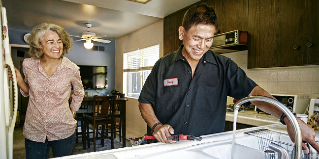 A handyman fixes a faucet as a homeowner looks over his shoulder in a kitchen.