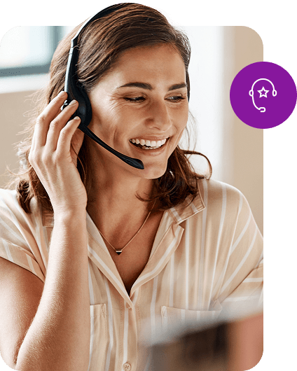 A smiling customer service representative wearing a headset assists someone over the phone while sitting at her computer.