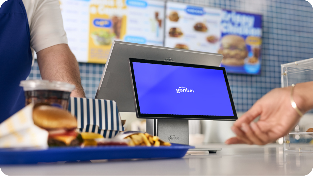 A Genius restaurant POS next to a tray of food in a QSR restaurant. Genius digital menu boards are in the background.