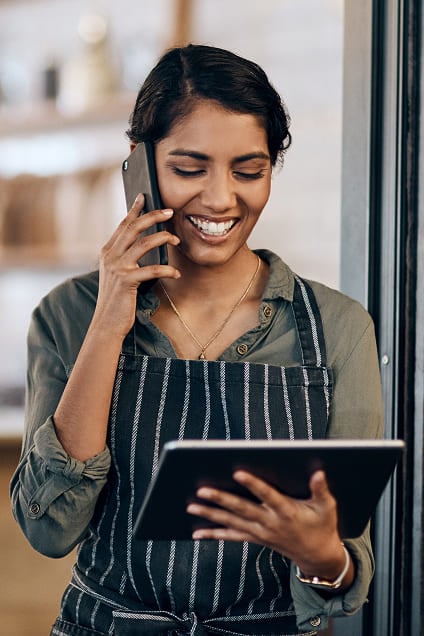Cafe worker smiling as she speaks on a phone while holding a digital tablet.