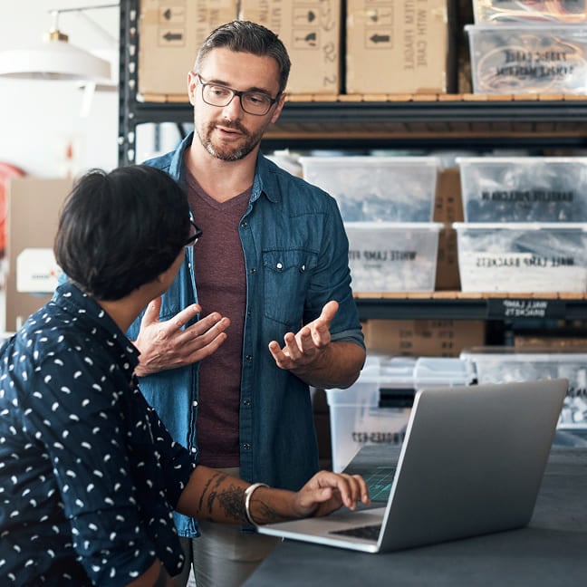 Man gesturing while talking to a colleague using a laptop in a warehouse.