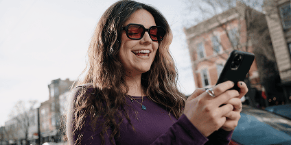 Smiling woman in sunglasses using a smartphone on a city street.