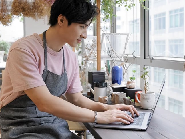 A café worker wearing an apron checks his laptop for payment solutions for his business.
