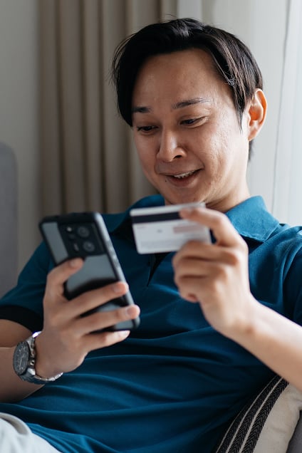 A man sits happily while entering the details of a card he’s holding into the mobile phone in his other hand.