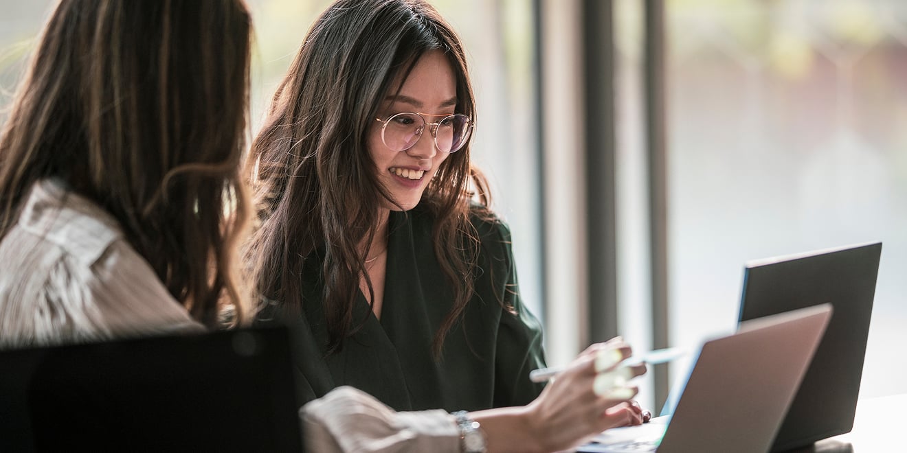 Two women sit at a table and work from two laptops while engaging in collaboration.