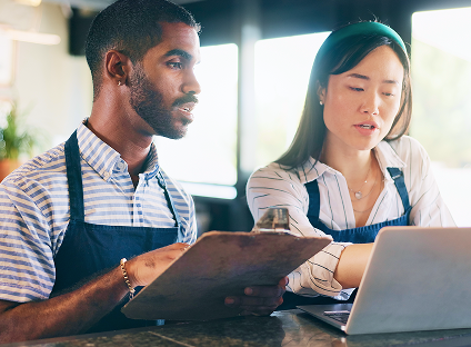 Two people wearing aprons work together over a laptop and clipboard.