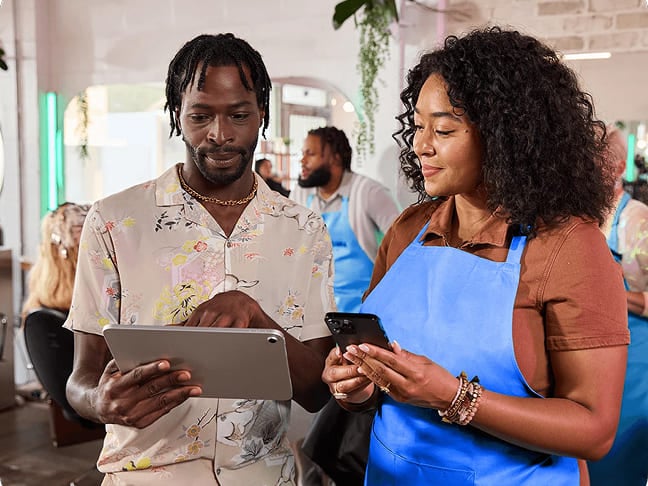 A man in a hair salon holds and points to a tablet while a female wearing an apron and holding a mobile device looks at the tablet with him.