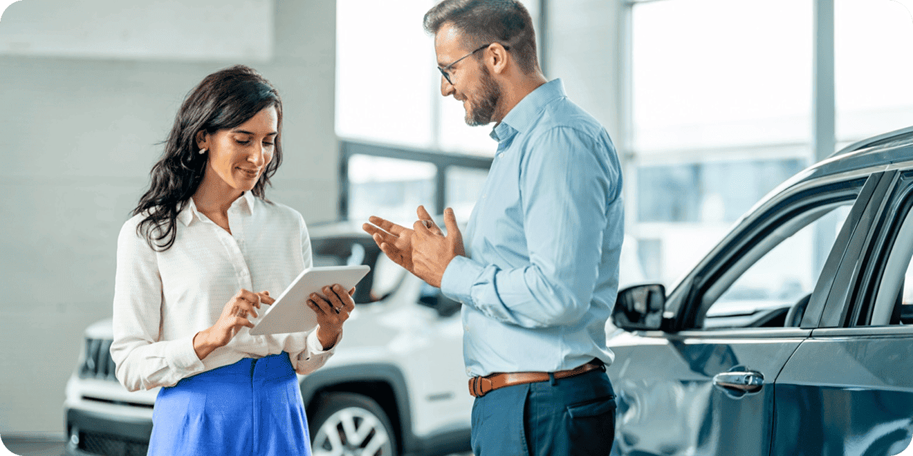 Woman with a tablet talking to a male customer in a car dealership.