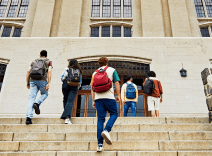 Five students with backpacks walk up wide stone steps toward the entrance of a large collegiate building.