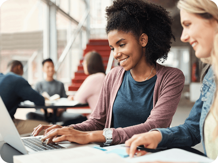 Two young women sit at a table in a common area of a university building and look at a laptop screen together.