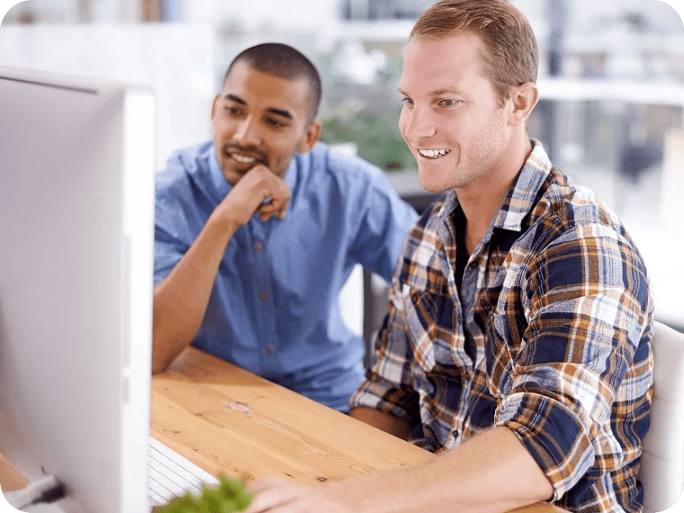 Two young men in button-down shirts sit at a desk and smile as they look at a computer screen together.