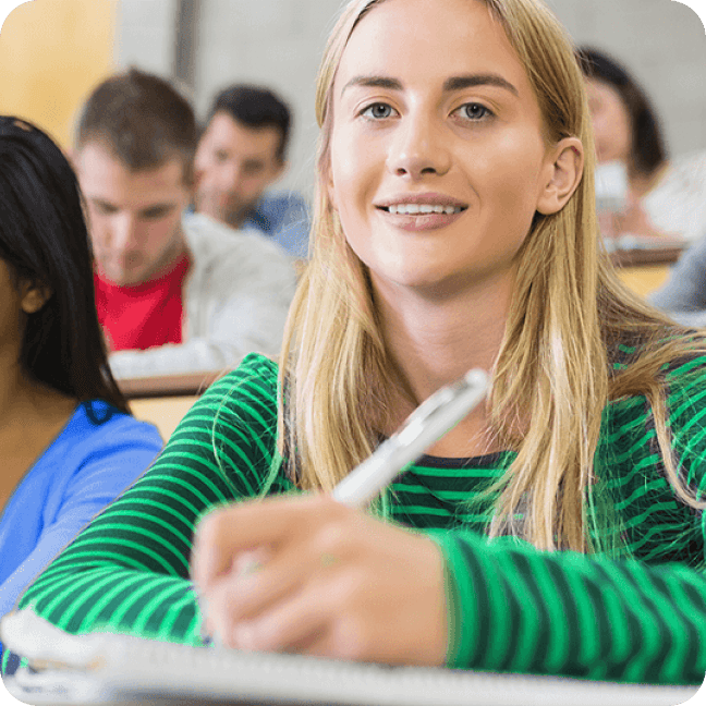 Une femme blonde souriante, vêtue d'une chemise rayée verte, prend des notes dans une salle de classe bondée.