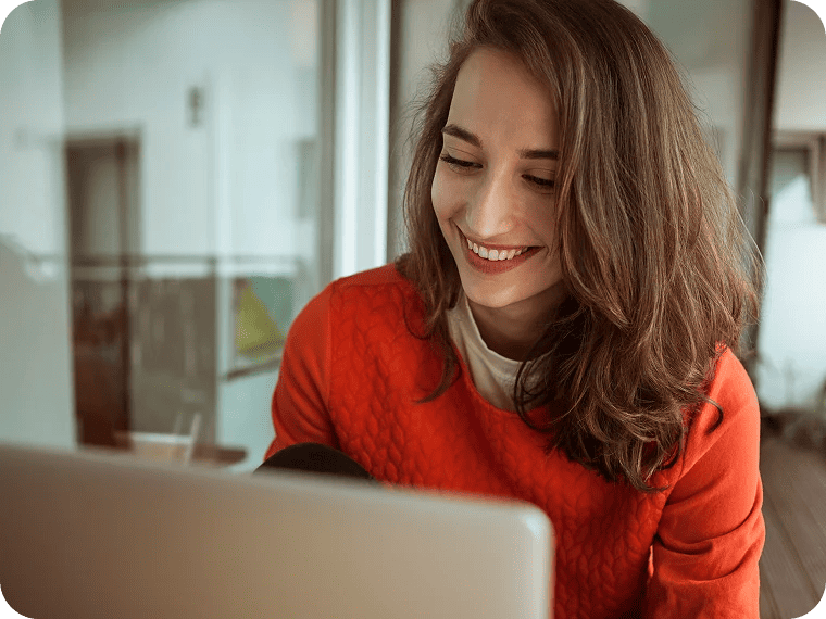 A young woman in a bright sweater smiles as she looks at a laptop screen.