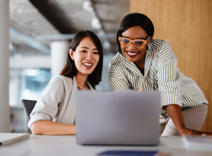 Two smiling colleagues look at a laptop screen together during a meeting.
