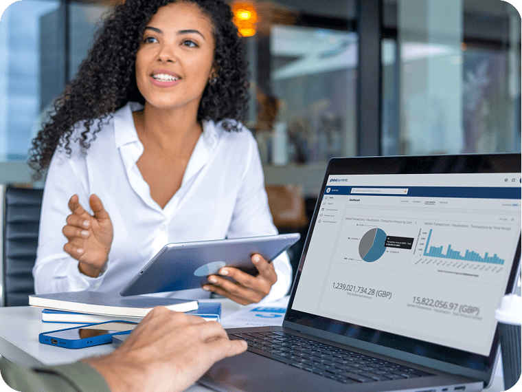 Woman gesturing with a tablet next to a laptop showing financial charts.