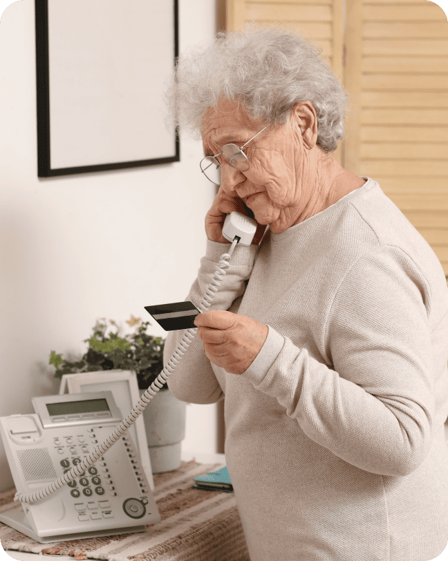 A standing woman speaks over a landline phone to make an IVR payment with the credit card in her hand.