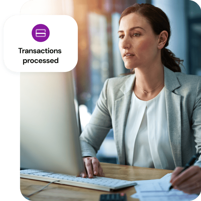 A woman sits at a table with a desktop computer and a graphic overlaying says transaction processed.
