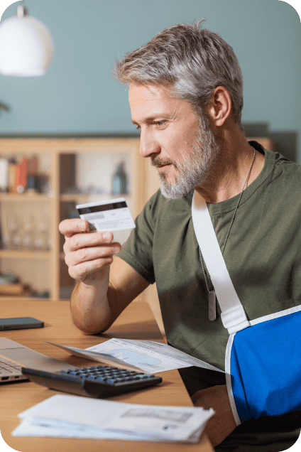 A man with a sling holds a credit card in his other hand and sits at a table with a laptop to make an online bill payment.