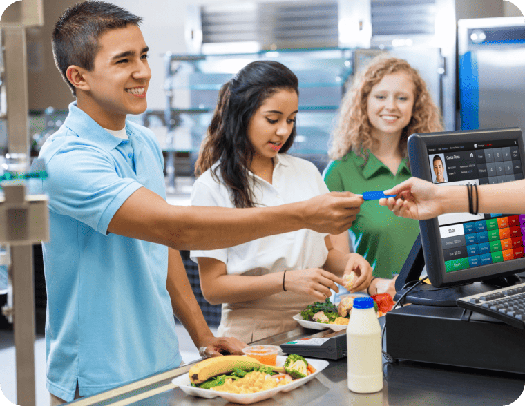 A student hands a card to a cashier at a cafeteria checkout counter, while two others stand behind with trays of food and drinks.