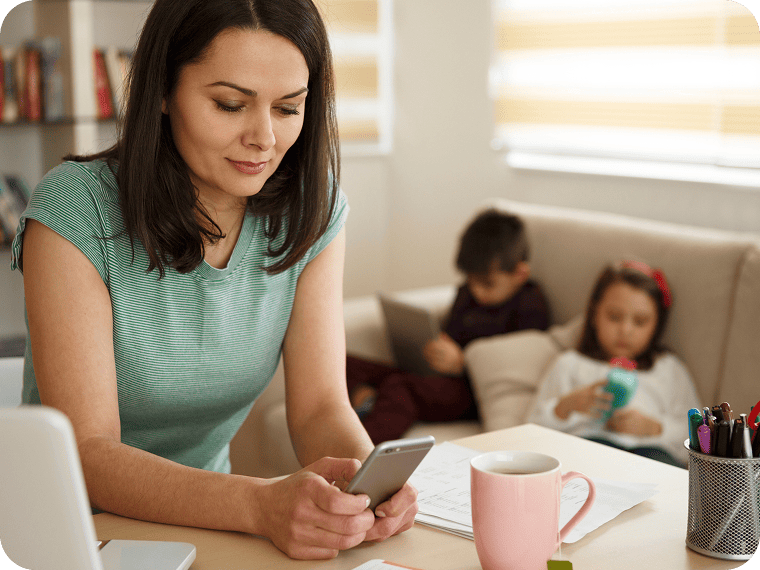 A woman smiles as she looks at her cell phone while sitting at her desk. Two kids relax on a couch beside her.