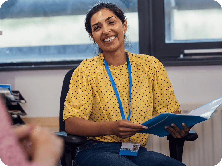 A smiling woman in a yellow, polka dot shirt is sitting in a chair and holding a folder.