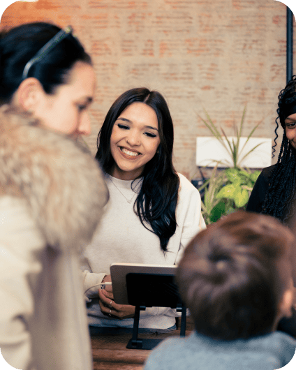 A smiling woman at a counter interacts with customers, including a child, in a cozy indoor setting with plants.