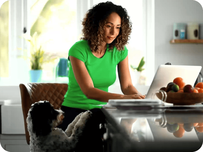 A woman in a green shirt works on a laptop at a kitchen counter while a black and white dog stands beside her, resting its paws on her leg.