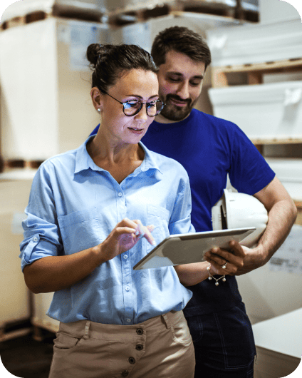 Woman using a tablet alongside a worker holding a hard hat in a warehouse.