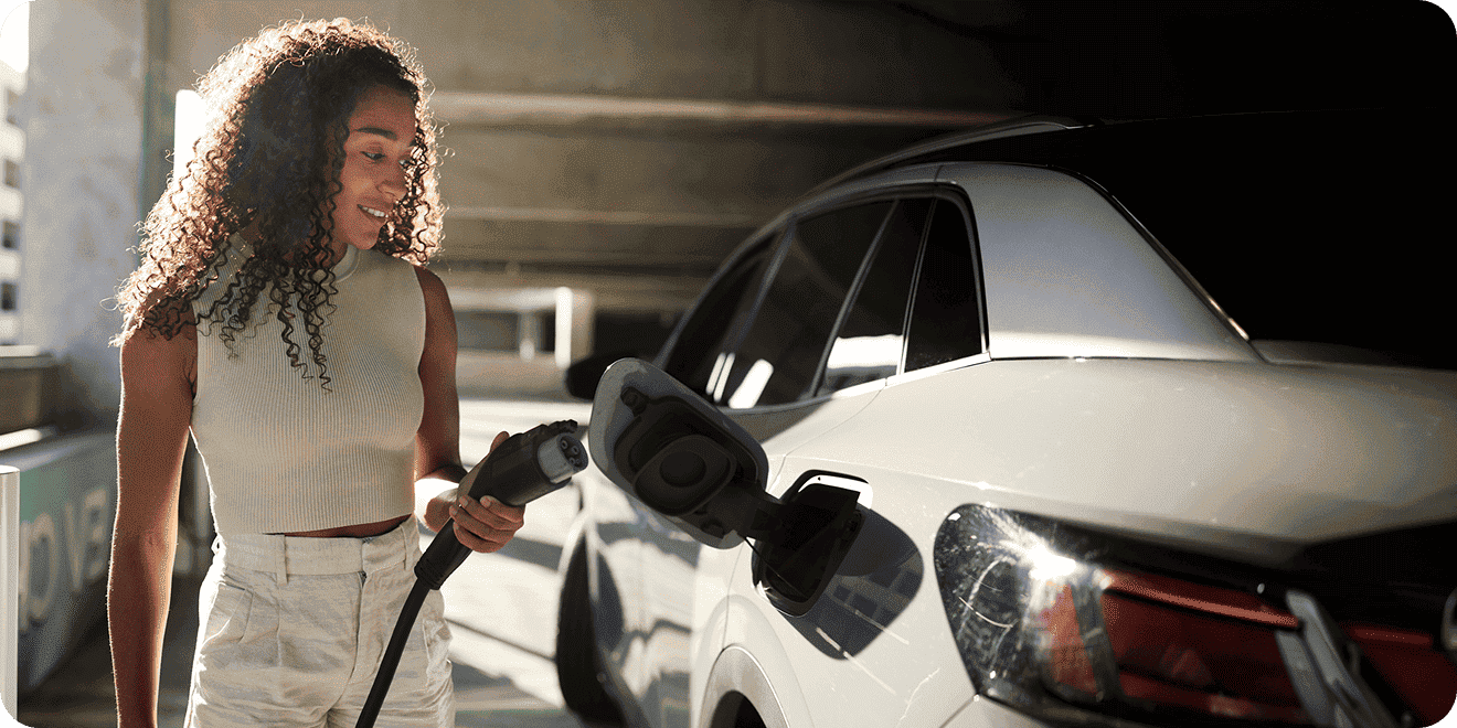 Woman with curly hair smiling while holding an EV charging cable by a white electric car.
