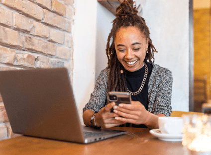 Woman with dreadlocks smiling while looking at her smartphone at a table.
