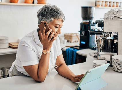 Woman talking on a mobile phone and using a tablet behind a café counter.