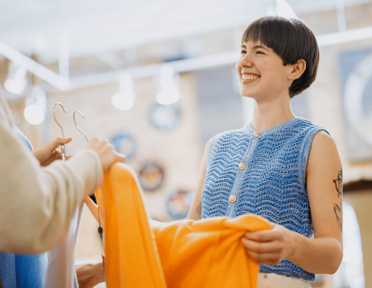 A smiling woman with short dark hair and a blue crocheted top looks at a bright orange garment held by another person in a clothing store.