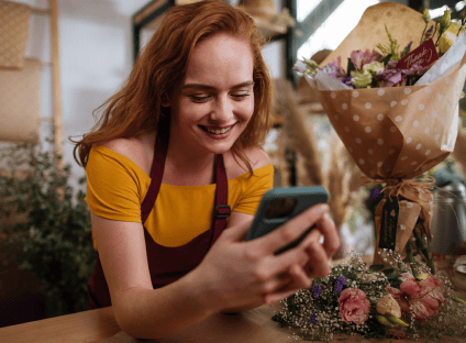 Smiling red-haired florist using a smartphone at a flower shop counter. 