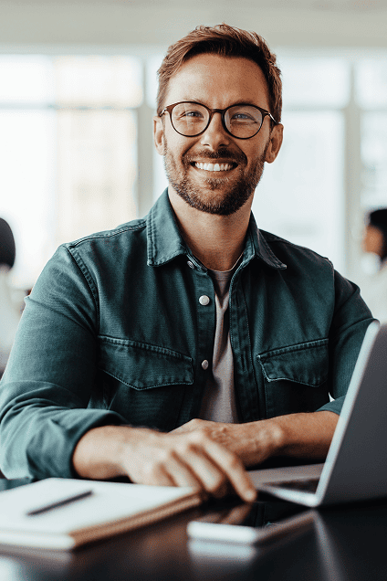A man in an open office sits in front of a laptop smiling at the camera